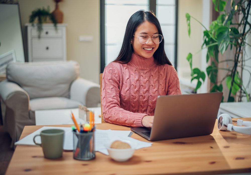 Young Vietnamese woman working in a home office and using a laptop. Flexible workspace and working time. Beautiful Asian business female or student typing an email or working on a project. Copy space.