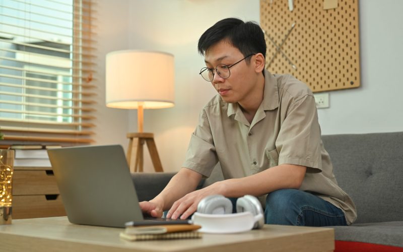 Young asian man sitting on couch and using laptop computer. Freelance, remote work and e-learning concept.